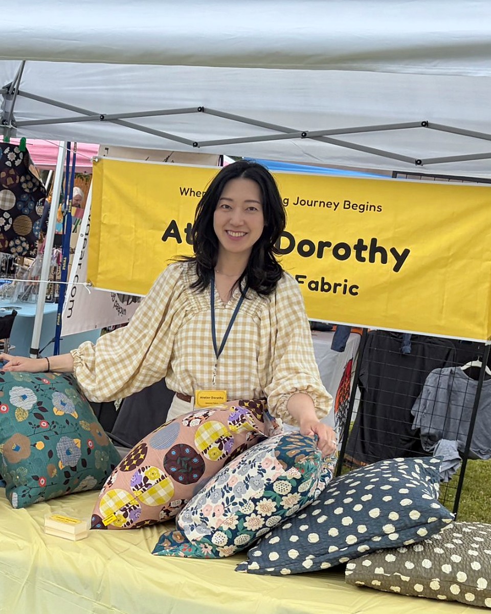 Mina, founder of Atelier Dorothy, smiling at her market booth surrounded by Japanese fabrics and handmade cushion covers